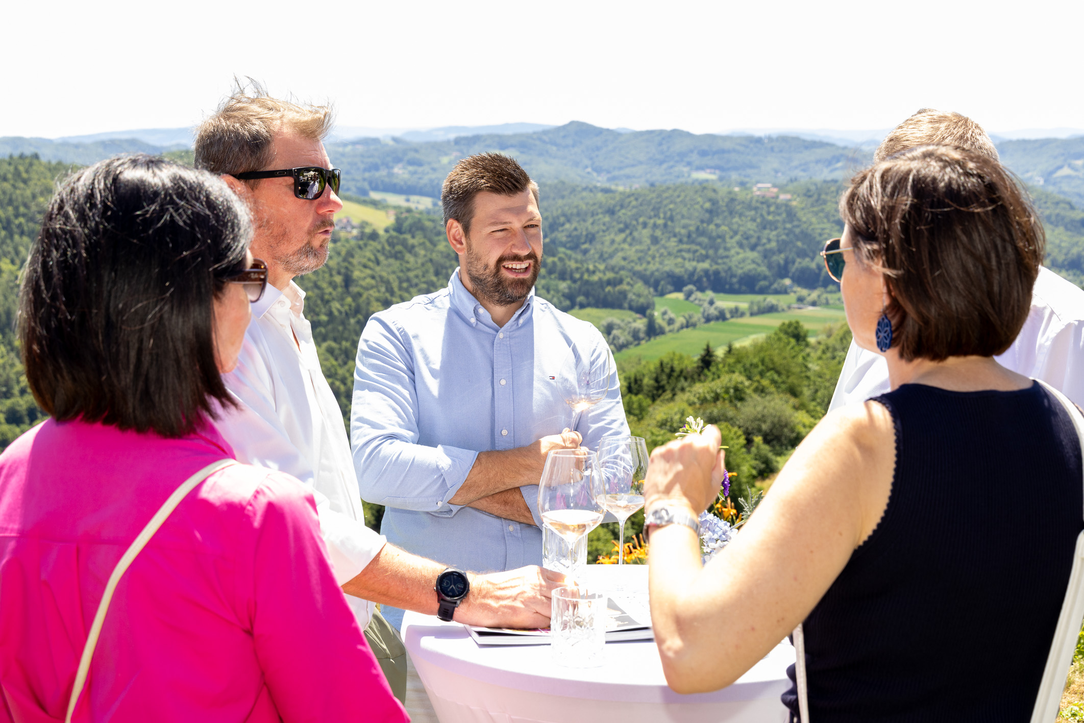 Vier Menschen stehen draußen um einen Tisch herum, unterhalten sich und lächeln, im Hintergrund sind malerische grüne Hügel und strahlender Sonnenschein zu sehen.