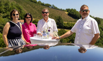 Vier Erwachsene mit Sonnenbrillen stehen um einen kleinen Tisch im Freien herum, auf dem Getränke stehen; im Hintergrund sind grüne Weinberge und der blaue Himmel zu sehen, die sich auf einer glänzenden Oberfläche spiegeln.
