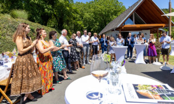 Eine Gruppe von Menschen steht im Freien, klatscht und lächelt bei einer Veranstaltung mit Getränken und Tischen bei sonnigem Wetter in der Nähe eines modernen Gebäudes.