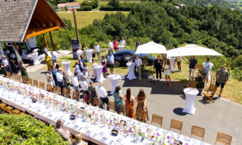 Eine Zusammenkunft im Freien an einem sonnigen Tag, bei der die Menschen an Cocktailtischen stehen, neben einem mit Blumen geschmückten langen Tisch, umgeben von grünen Hügeln.