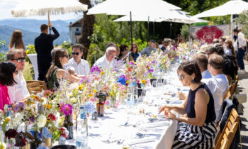 Menschen sitzen im Freien an einem langen Tisch, der mit bunten Blumen geschmückt ist, und genießen an einem sonnigen Tag unter weißen Sonnenschirmen eine Mahlzeit.
