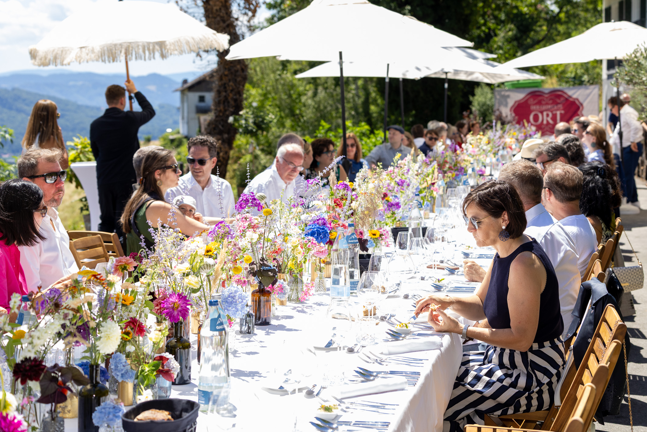 Menschen sitzen im Freien an einem langen Tisch, der mit bunten Blumen geschmückt ist, und genießen an einem sonnigen Tag unter weißen Sonnenschirmen eine Mahlzeit.