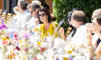 Eine Gruppe von Menschen sitzt an einem mit bunten Blumen geschmückten Tisch im Freien und genießt das Essen und die Unterhaltung im strahlenden Sonnenschein.