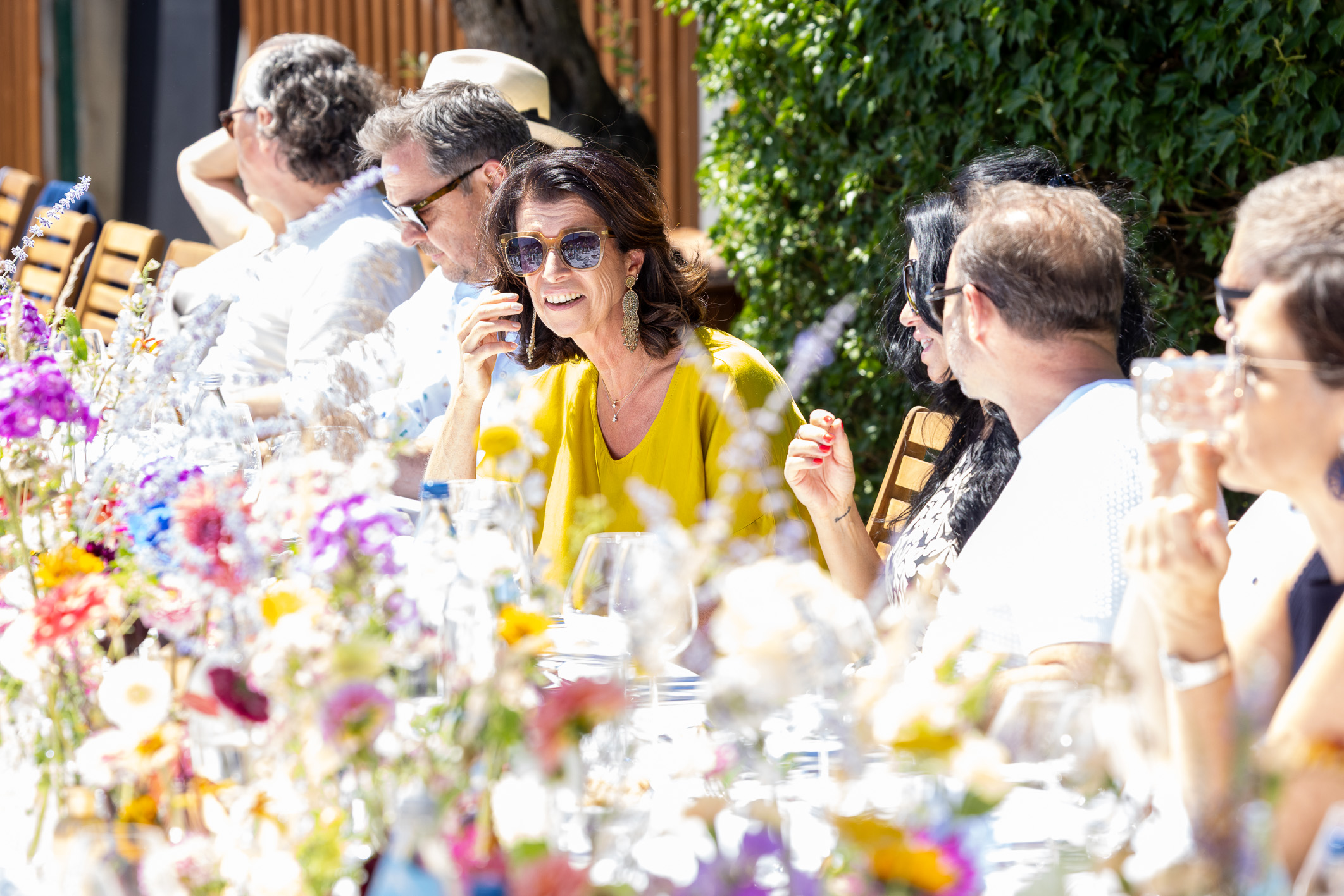 Eine Gruppe von Menschen sitzt an einem mit bunten Blumen geschmückten Tisch im Freien und genießt das Essen und die Unterhaltung im strahlenden Sonnenschein.