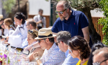Eine Gruppe von Menschen sitzt im Sonnenlicht an einem langen Tisch im Freien, genießt gemeinsam Wein und Essen, während ein Mann in einem blauen Polohemd steht und sich mit den Gästen unterhält.