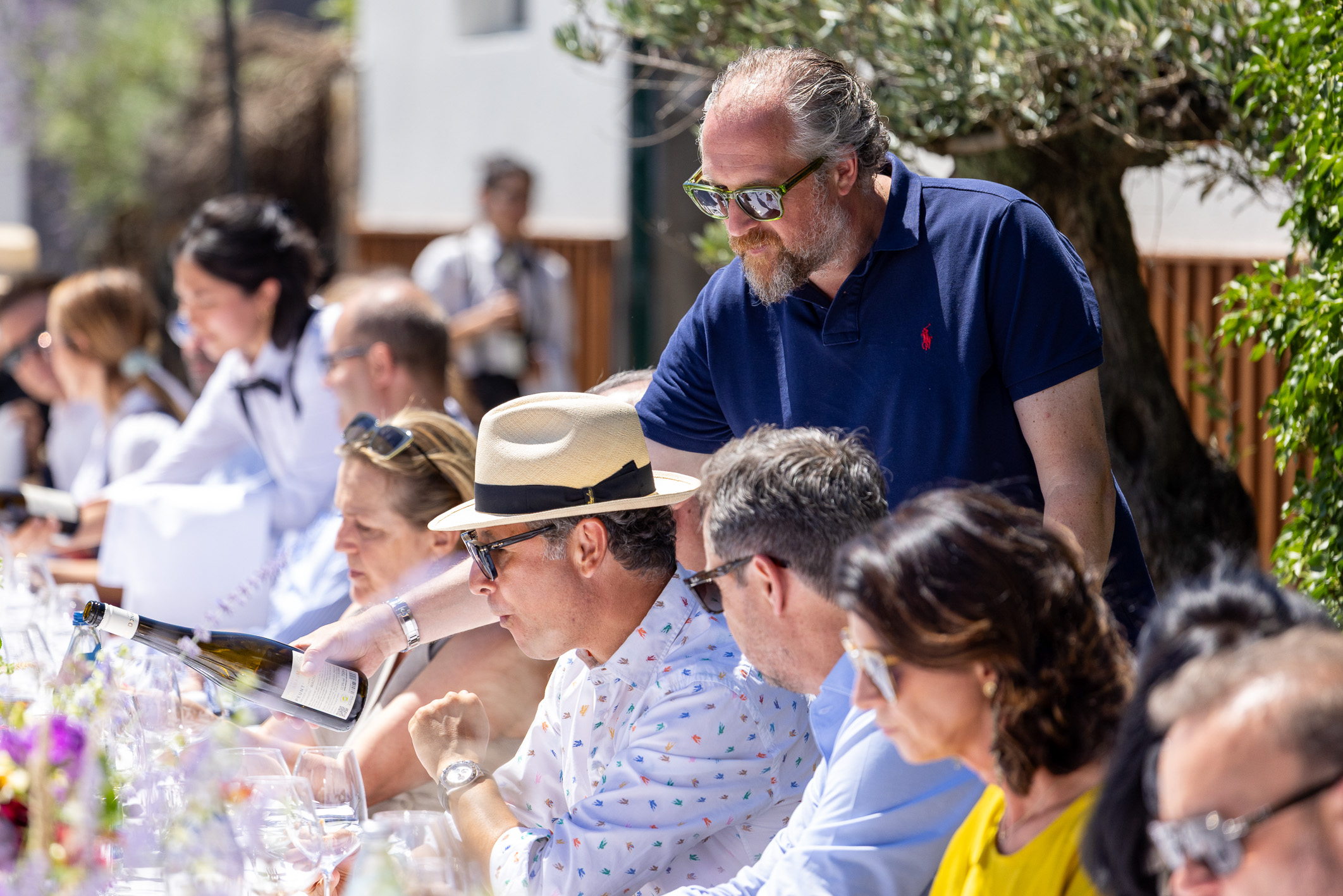Eine Gruppe von Menschen sitzt im Sonnenlicht an einem langen Tisch im Freien, genießt gemeinsam Wein und Essen, während ein Mann in einem blauen Polohemd steht und sich mit den Gästen unterhält.
