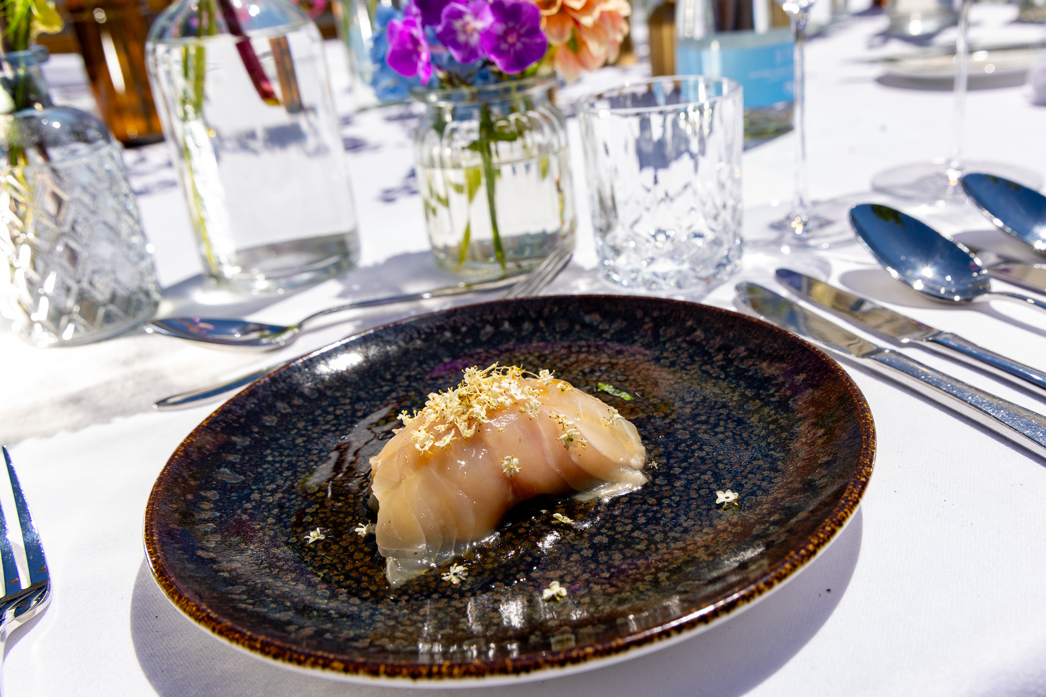 Ein Sushi-Gericht, garniert mit Goldflocken auf einem dunklen Teller, serviert auf einem eleganten Tisch mit Blumen, Gläsern und Besteck.