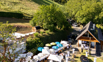 Auf einem Weinberghang findet „Der gedeckte Ort, Band 2“ statt – mit Tischen im Freien, Sonnenschirmen und Gästen, die sich an einem sonnigen Tag vor einem Holzgebäude versammelt haben.