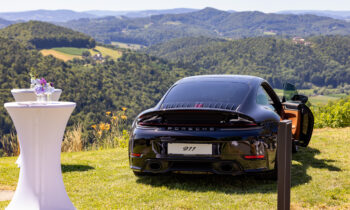 Ein schwarzer Porsche 911 steht auf einer Rasenfläche, im Hintergrund sind malerische Hügel zu sehen; daneben steht ein Cocktailtisch mit Blumen.