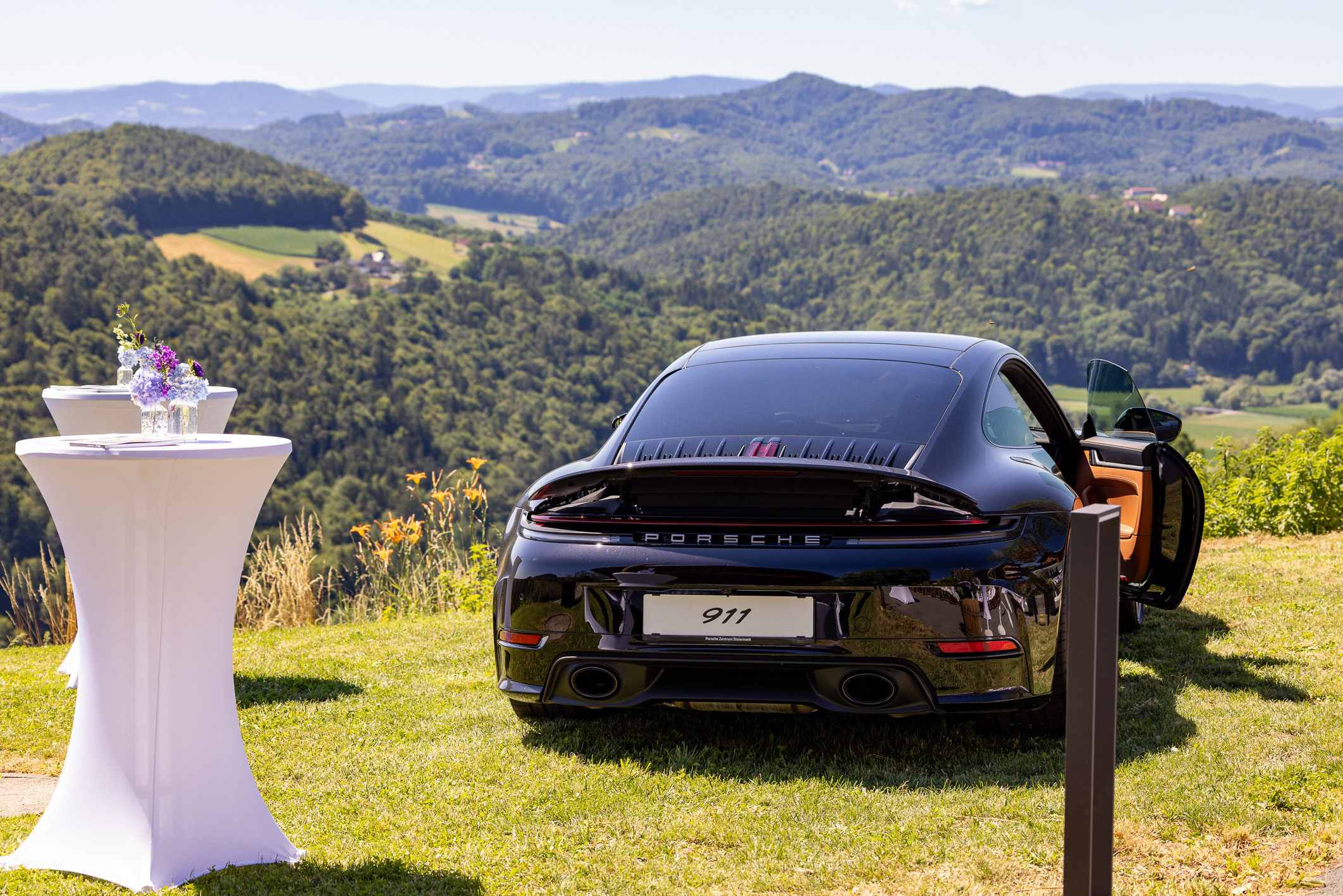 Ein schwarzer Porsche 911 steht auf einer Rasenfläche, im Hintergrund sind malerische Hügel zu sehen; daneben steht ein Cocktailtisch mit Blumen.