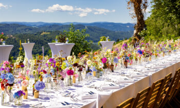 Ein langer Esstisch im Freien mit weißer Tischdecke, bunten Blumenarrangements, Holzstühlen und einem malerischen Blick auf die Berge unter strahlend blauem Himmel.