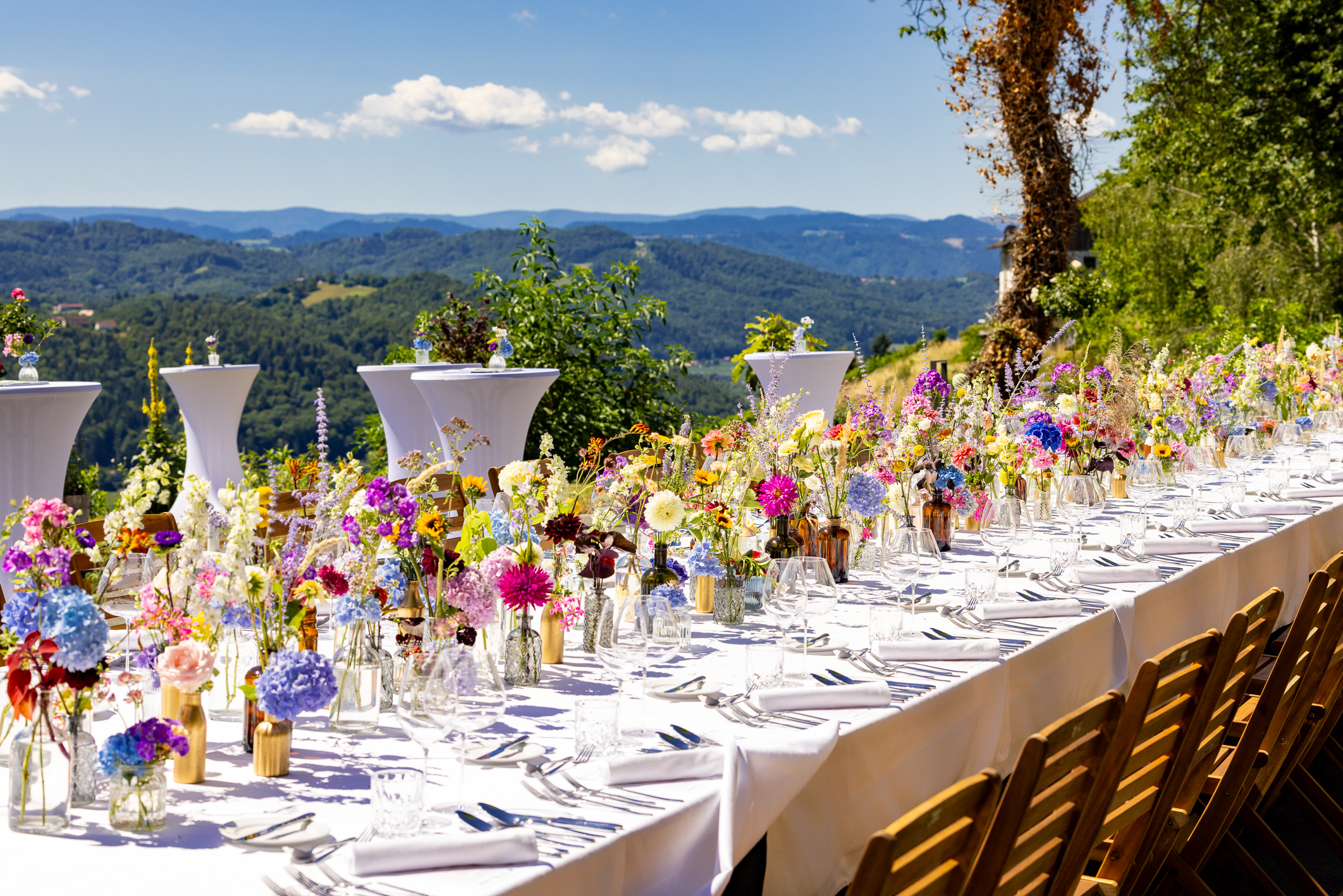 Ein langer Esstisch im Freien mit weißer Tischdecke, bunten Blumenarrangements, Holzstühlen und einem malerischen Blick auf die Berge unter strahlend blauem Himmel.