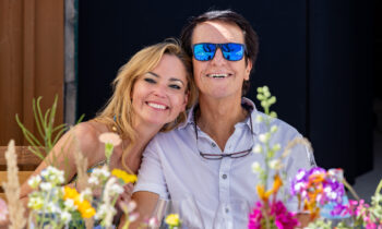 Eine lächelnde Frau und ein Mann sitzen an einem mit bunten Blumen geschmückten Tisch im Freien an einem sonnigen Tag dicht beieinander.
