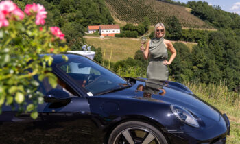 Eine Frau in einem grünen Kleid hält ein Glas Wein neben einem schwarzen Sportwagen in der Hand – „Der gedeckte Ort, Band 2“ spielt inmitten von Hügeln, Weinbergen und einem Haus.