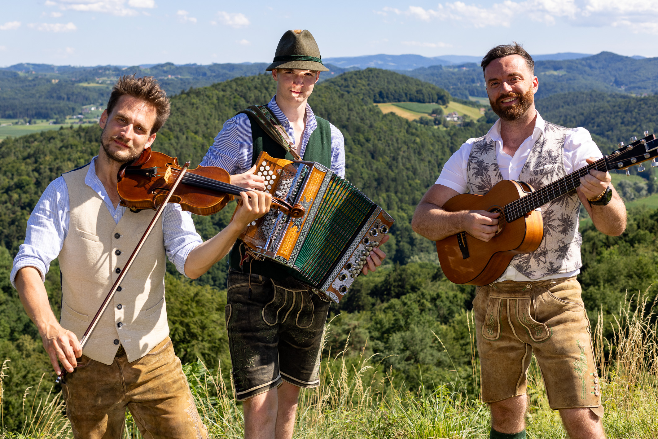 Drei Männer in traditioneller bayerischer Tracht spielen im Freien Geige, Akkordeon und Gitarre, vor dem Hintergrund grüner Hügel und blauem Himmel.