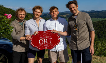 Vier Männer stehen an einem sonnigen Tag im Freien und halten ein rotes Schild mit der Aufschrift „Der Gedeckte Ort 2026“ in den Händen; im Hintergrund sind grüne Hügel und Bäume zu sehen.