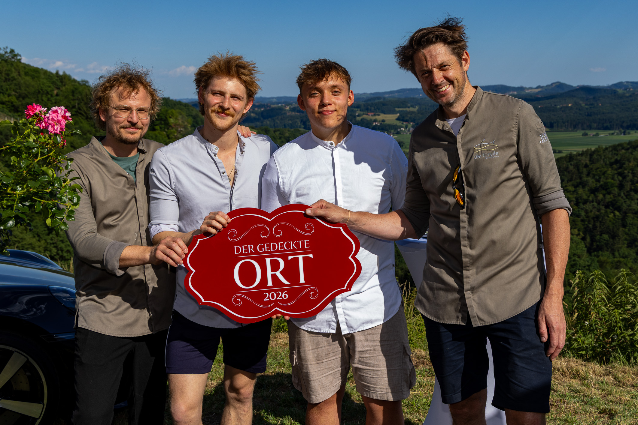 Vier Männer stehen an einem sonnigen Tag im Freien und halten ein rotes Schild mit der Aufschrift „Der Gedeckte Ort 2026“ in den Händen; im Hintergrund sind grüne Hügel und Bäume zu sehen.