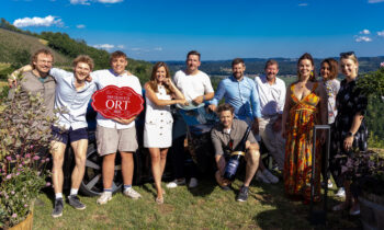 Eine Gruppe von elf Personen posiert an einem sonnigen Tag im Freien, lächelt vor malerischen Hügeln und blauem Himmel und hält ein rotes Schild sowie eine Weinflasche in den Händen.