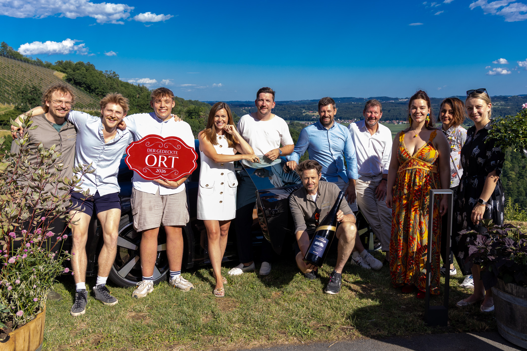Eine Gruppe von elf Personen posiert an einem sonnigen Tag im Freien, lächelt vor malerischen Hügeln und blauem Himmel und hält ein rotes Schild sowie eine Weinflasche in den Händen.