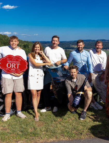 Eine Gruppe von elf Personen, die an einem sonnigen Tag im Freien lächeln und auf einer Wiese stehen, im Hintergrund sind Hügel und der blaue Himmel zu sehen. Einige halten ein rotes Schild und einen großen Schlüssel in der Hand.