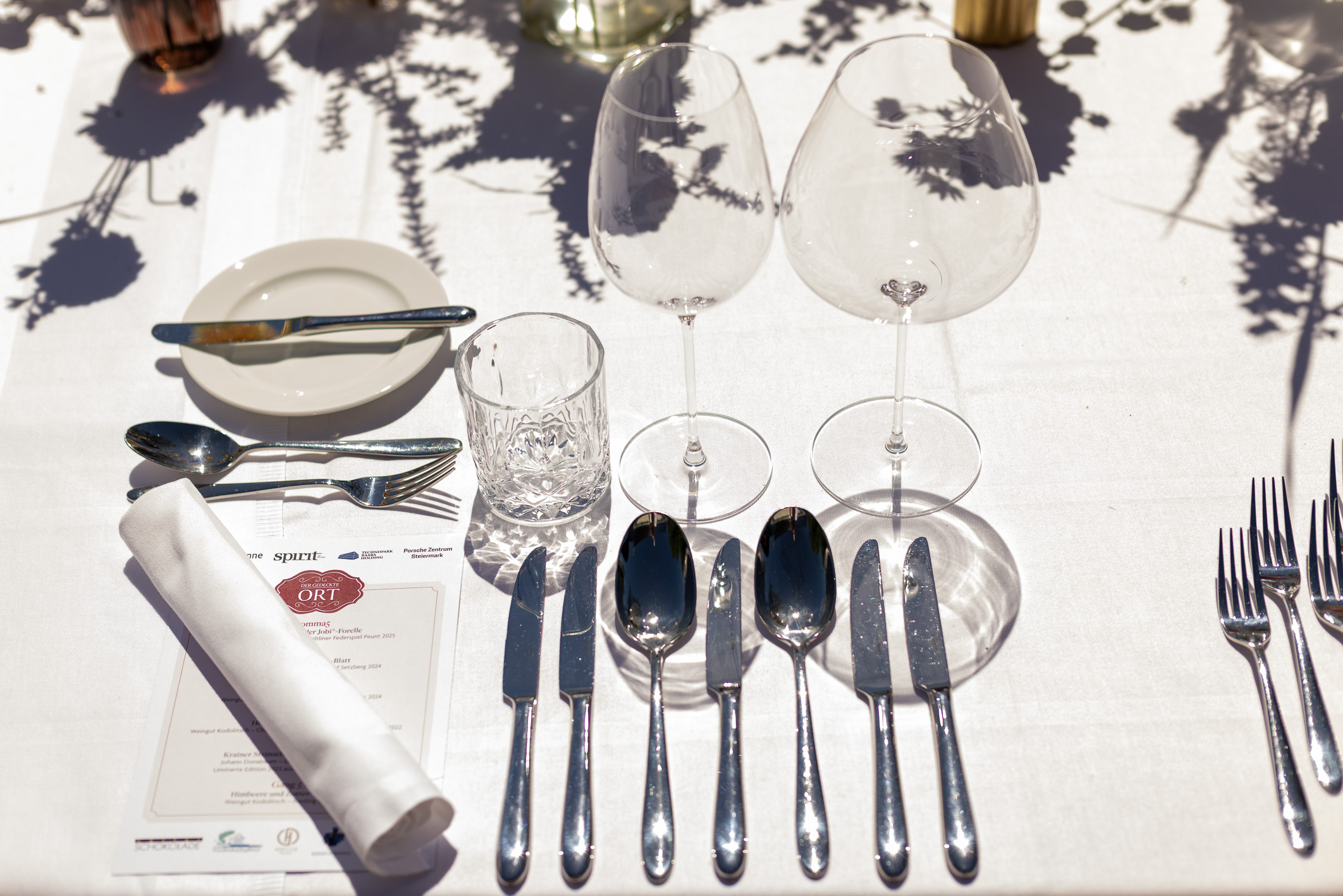 Elegante Tischdekoration mit mehreren Gläsern, Messern, Gabeln, einer Serviette, einer Speisekarte und einem kleinen Teller auf einer weißen Tischdecke im Sonnenlicht.