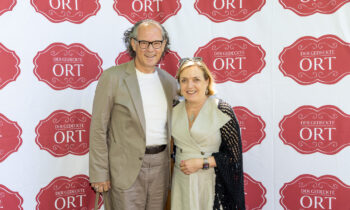 Ein Mann und eine Frau lächeln, während sie gemeinsam vor einem Hintergrund stehen, auf dem sich das Logo „Der Gedeckte Ort“ wiederholt.