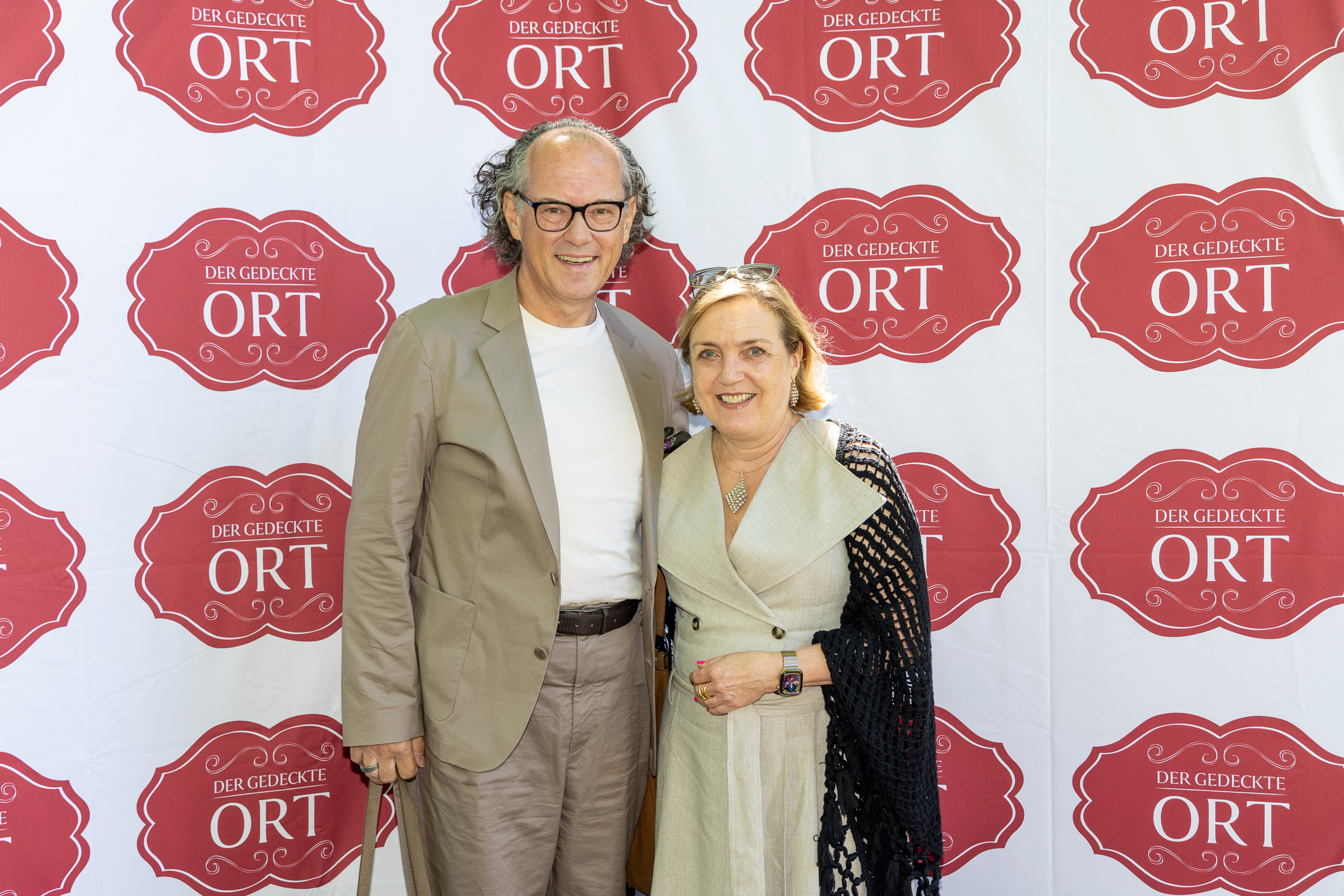 Ein Mann und eine Frau lächeln, während sie gemeinsam vor einem Hintergrund stehen, auf dem sich das Logo „Der Gedeckte Ort“ wiederholt.