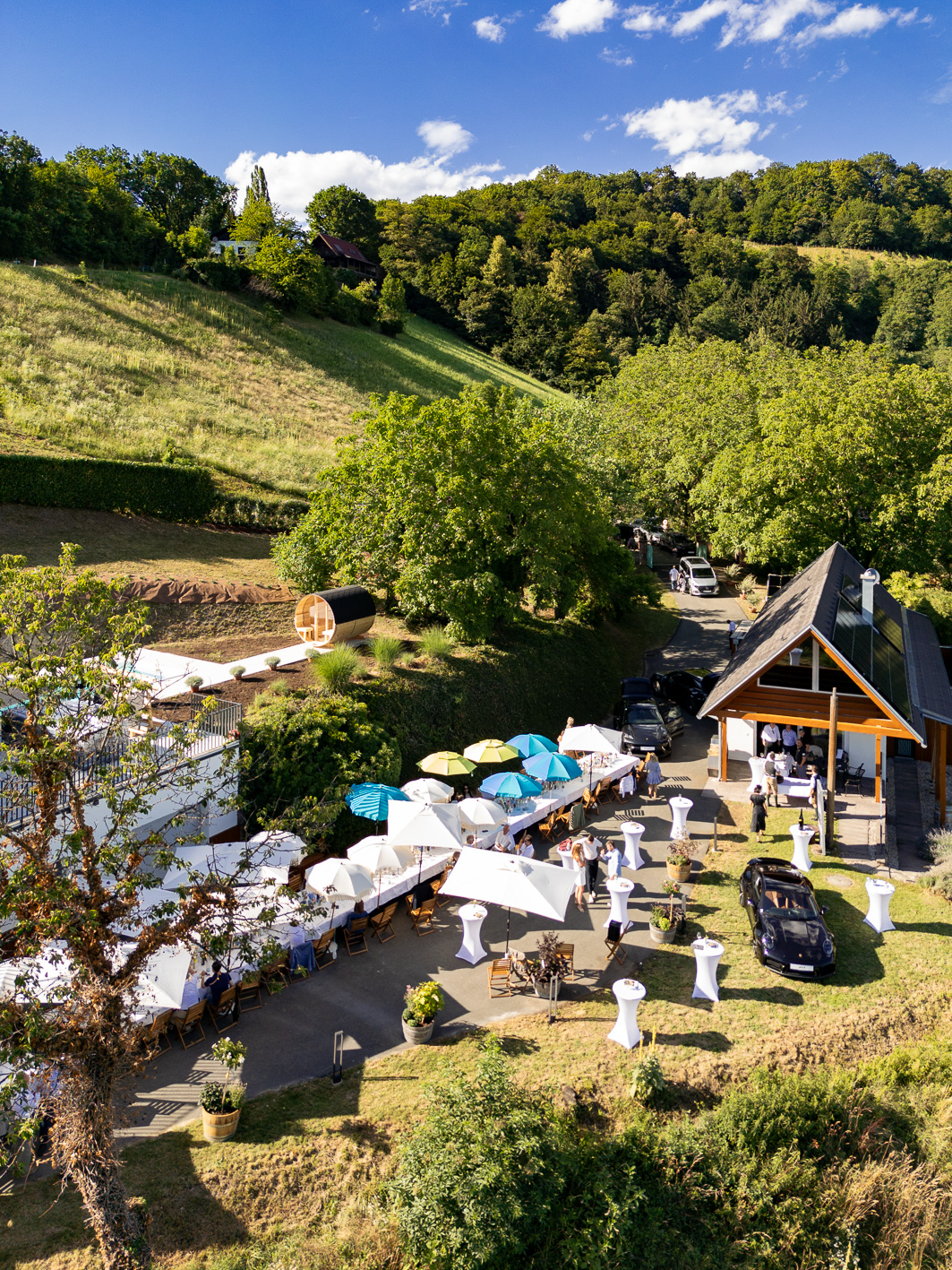 Auf einem Weinberghang findet „Der gedeckte Ort, Band 2“ statt – mit Tischen im Freien, Sonnenschirmen und Gästen, die sich an einem sonnigen Tag vor einem Holzgebäude versammelt haben.