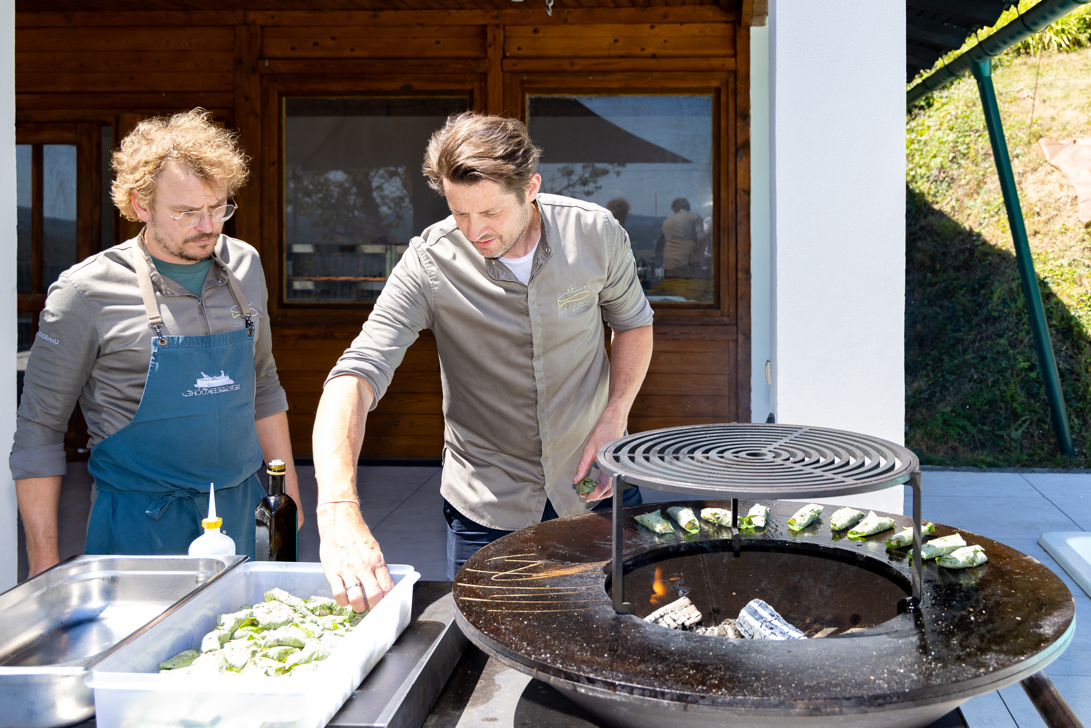Zwei Männer kochen im Freien; einer legt Lebensmittel auf den Grill, während der andere, der eine Schürze trägt, neben einem Tablett mit vorbereiteten Zutaten steht und zusieht.