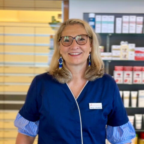 Eine lächelnde Frau mit Brille in einer blauen Uniform steht in einer Apotheke, im Hintergrund sind Regale mit Produkten zu sehen.