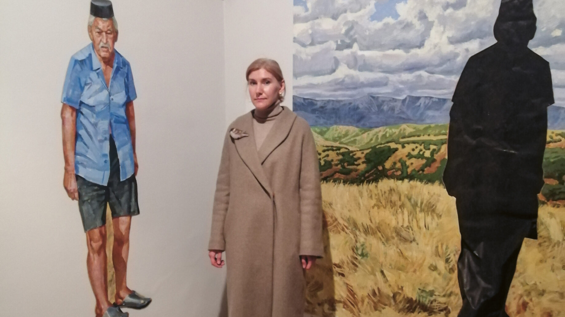 In einem Galerieraum steht eine Frau in einem hellbraunen Mantel zwischen einem Wandgemälde, das einen Mann zeigt, und einem großen Gemälde mit einer schwarzen Silhouette.