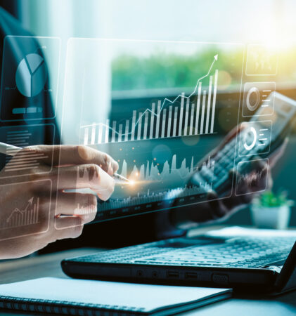 Eine Person, die auf einem Laptop mit einem Stylus virtuelle Finanzdiagramme analysiert, umgeben von Charts und Sonnenlicht, das durch ein Fenster fällt.