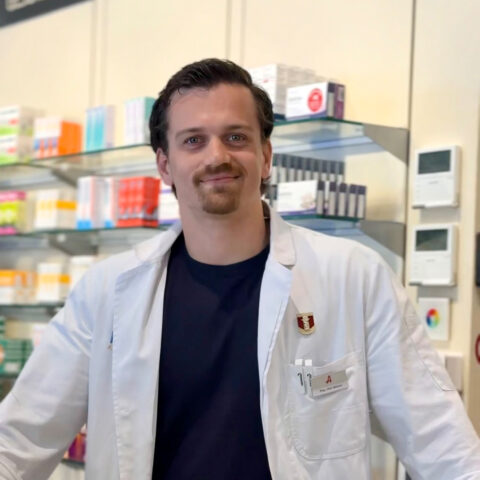 Ein Apotheker in einem weißen Kittel steht lächelnd in einer Apotheke, hinter ihm stehen Regale mit Medikamenten und anderen Produkten.