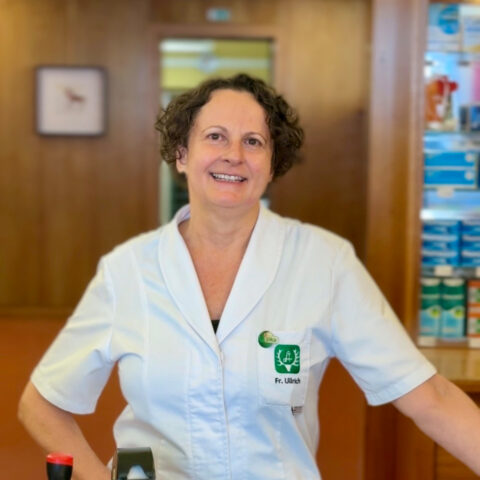 Eine Frau in einem weißen Kittel steht lächelnd in einer Apotheke, im Hintergrund sind Regale mit Produkten zu sehen.
