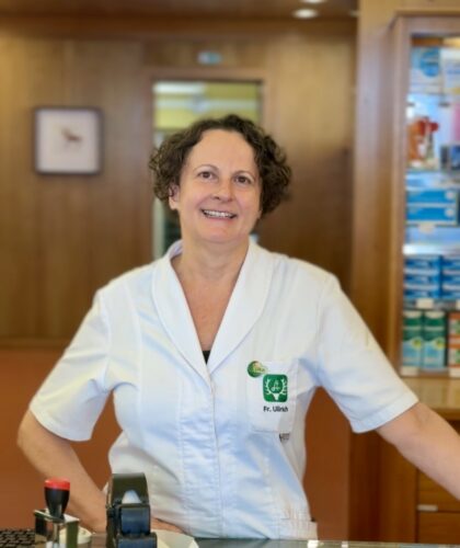 Karin Ullrich aus Deutschlandsberg strahlt als Apothekenstar in ihrem weißen Kittel, umgeben von Medikamenten in ihrer modernen Apotheke.