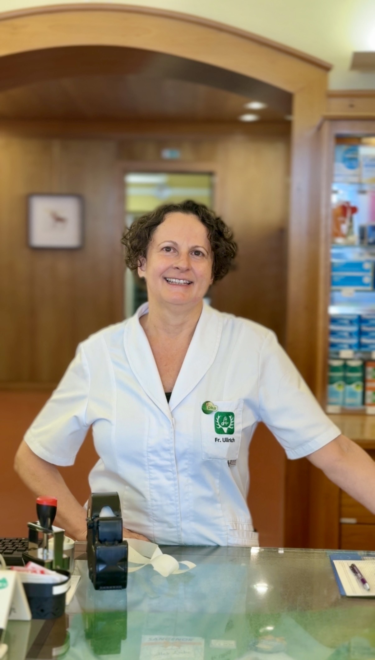Karin Ullrich aus Deutschlandsberg strahlt als Apothekenstar in ihrem weißen Kittel, umgeben von Medikamenten in ihrer modernen Apotheke.