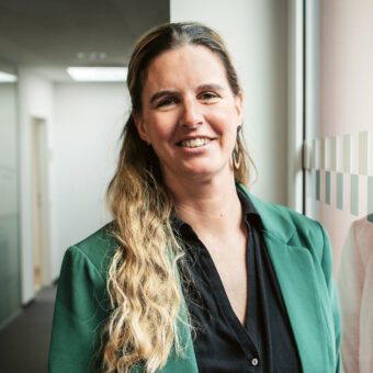 Eine Frau mit langen, welligen Haaren, die einen grünen Blazer und ein schwarzes Hemd trägt, steht lächelnd an einem Fenster in einem modernen Büroflur.