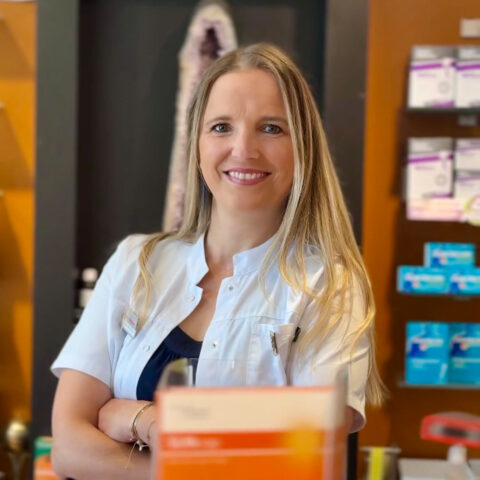 In einer Apotheke steht eine Frau in einem weißen Kittel lächelnd mit verschränkten Armen da; hinter ihr sind Medikamentenschachteln in den Regalen zu sehen.