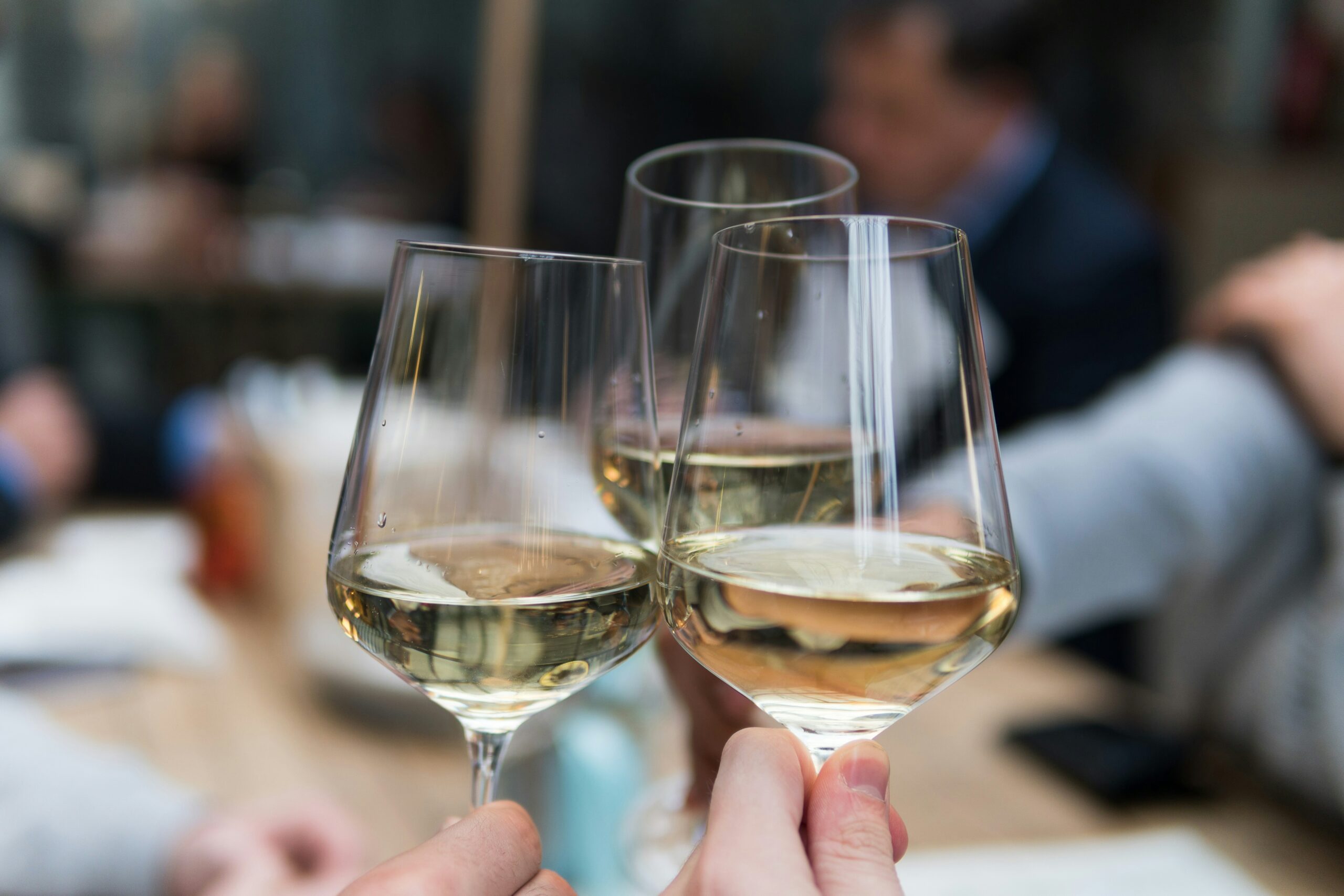 Drei Personen, die an einem Tisch mit Weißweingläsern anstoßen, im Hintergrund sind andere Gäste verschwommen zu sehen.