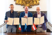 Drei Männer in Anzügen halten unter den Flaggen Italiens, der EU und Österreichs unterzeichnete Dokumente in den Händen und feiern vor einem prunkvollen Hintergrund die Vergabe der WKO-Steiermark-Forschungsstipendien 2026.
