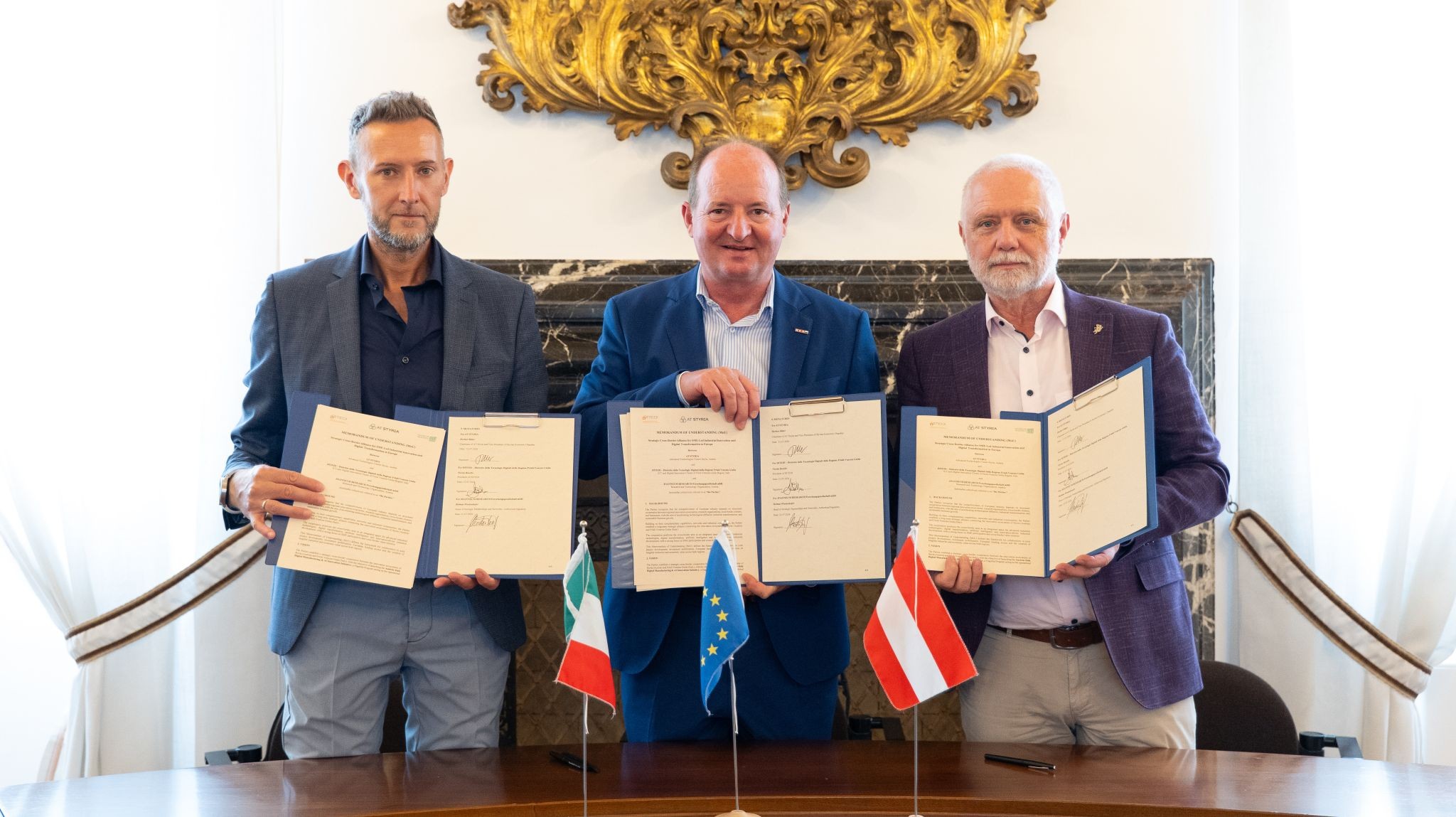 Drei Männer in Anzügen halten unter den Flaggen Italiens, der EU und Österreichs unterzeichnete Dokumente in den Händen und feiern vor einem prunkvollen Hintergrund die Vergabe der WKO-Steiermark-Forschungsstipendien 2026.