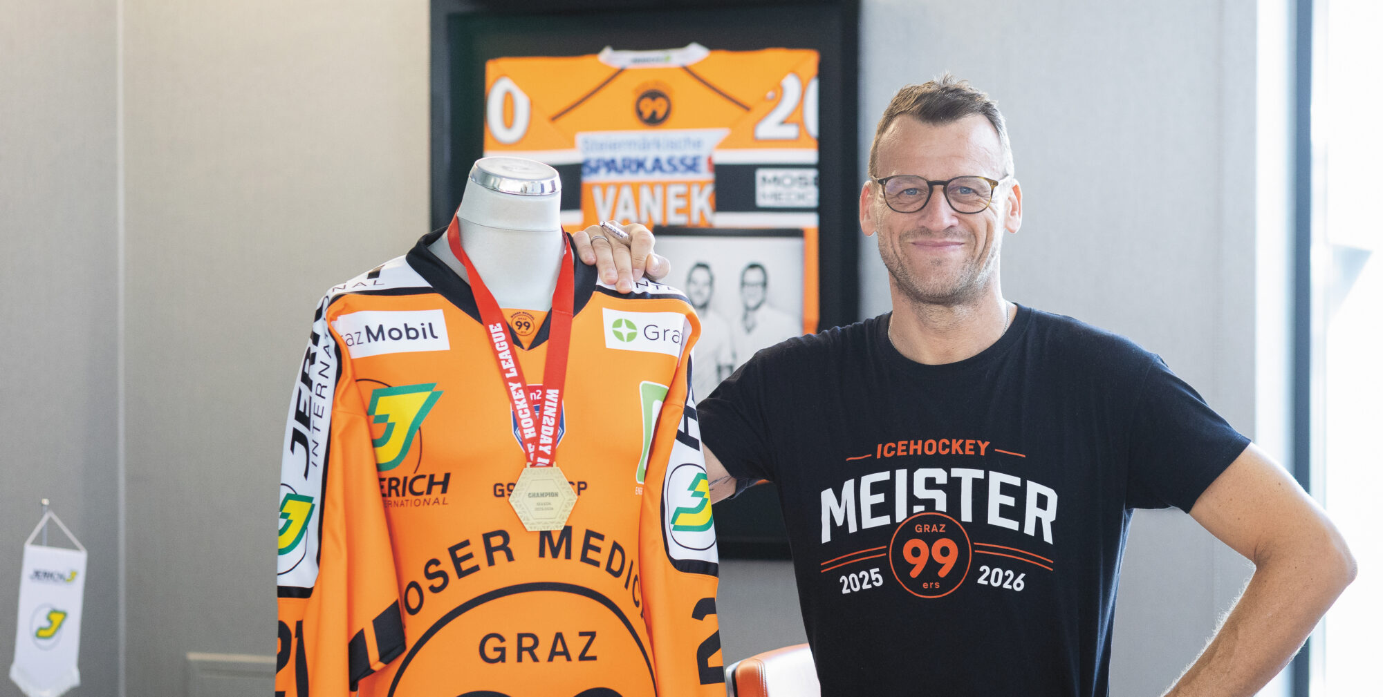 Ein lächelnder Mann mit Brille steht neben einem orangefarbenen Eishockeytrikot, das an einer Schaufensterpuppe mit einer Medaille hängt, und posiert in einem modernen Raum mit Erinnerungsstücken des Teams.
