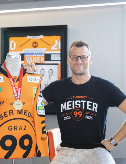 Ein lächelnder Mann mit Brille steht neben einem orangefarbenen Eishockeytrikot, das an einer Schaufensterpuppe mit einer Medaille hängt, und posiert in einem modernen Raum mit Erinnerungsstücken des Teams.