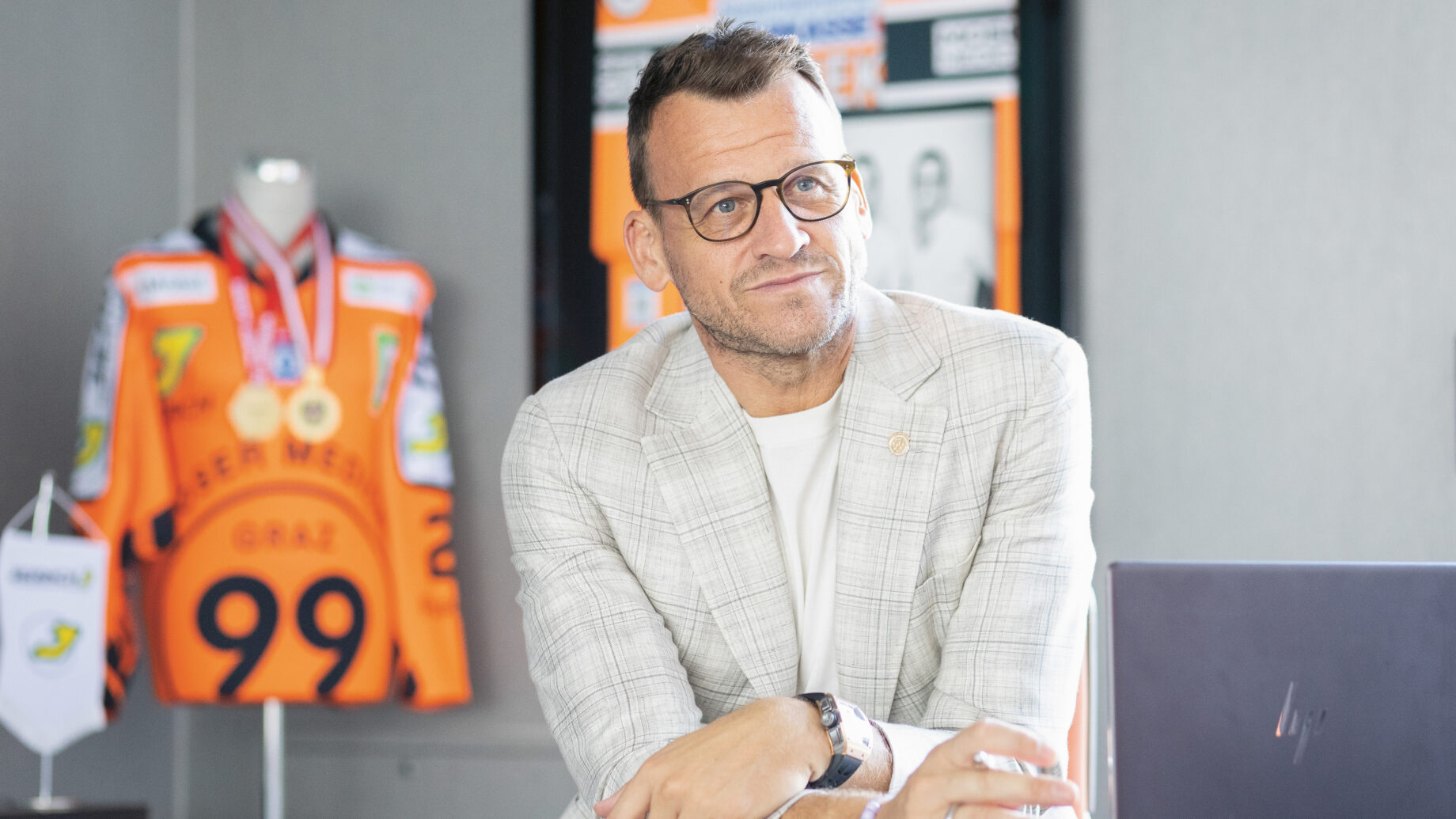 Ein Mann mit Brille, der einen hellen karierten Blazer trägt, sitzt an einem Schreibtisch mit einem Laptop; im Hintergrund sind ein Sporttrikot und Medaillen zu sehen.
