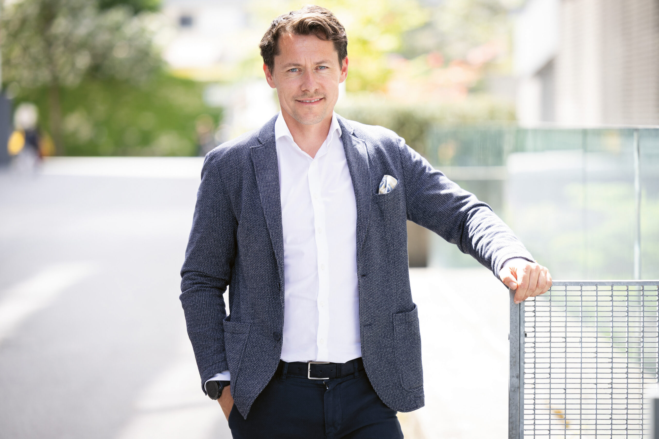 Ein Mann in einem blauen Blazer und einem weißen Hemd steht lächelnd im Freien, eine Hand in der Hosentasche und einen Arm auf einem Metallzaun abgestützt.
