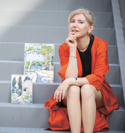 Eine Frau in einem roten Mantel sitzt lächelnd auf einer Treppe, neben ihr stehen zwei Landschaftsbilder.