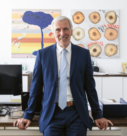 Ein Mann in einem blauen Anzug steht lächelnd in einem hellen Büro und lehnt sich an einen Schreibtisch, hinter dem sich Computer, Bücherregale und farbenfrohe Kunstwerke an der Wand befinden.