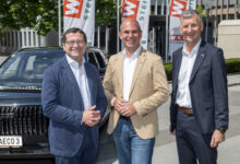 Drei Männer in Geschäftskleidung lächeln im Freien vor einem schwarzen Auto auf der Fahrzeugmarkt Steiermark 2026, hinter ihnen sind moderne Büros und Firmenfahnen zu sehen.