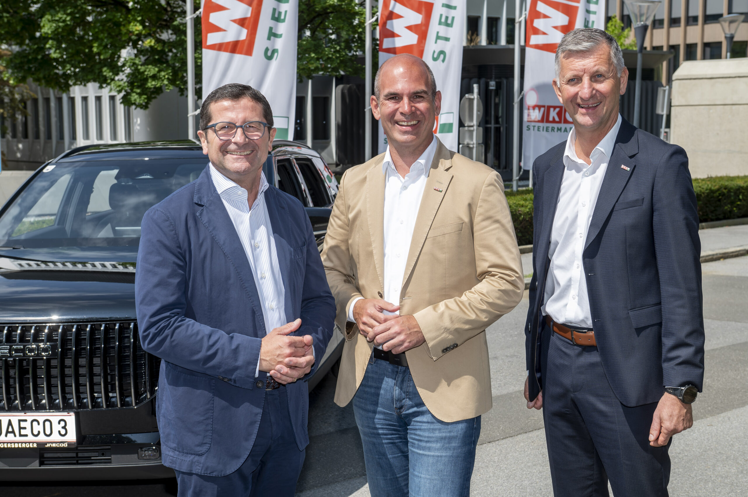 Drei Männer in Geschäftskleidung lächeln im Freien vor einem schwarzen Auto auf der Fahrzeugmarkt Steiermark 2026, hinter ihnen sind moderne Büros und Firmenfahnen zu sehen.