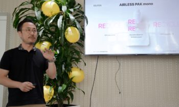 Auf dem Raiffeisen-Agrarsymposium präsentiert ein Mann „AIRLESS PAK mono“ neben einem Fernseher, auf dem „Reduce, Reuse, Recycle“ zu sehen ist, zusammen mit gelben Luftballons und einer Pflanze.